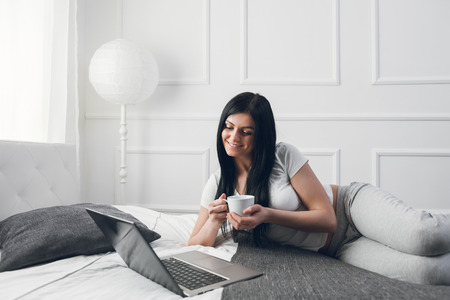 Relaxing on the bed. Beautiful young woman enjoying a cup of coffee and using her laptopの写真素材