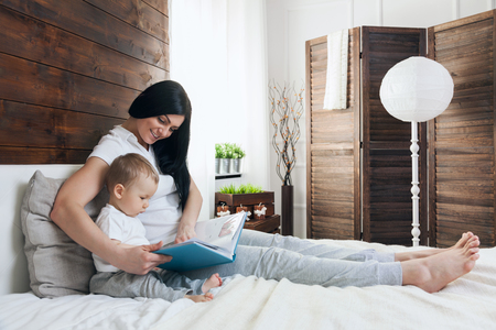 Child education. Happy mother with her toddler sitting on the bed and reading a book togetherの写真素材