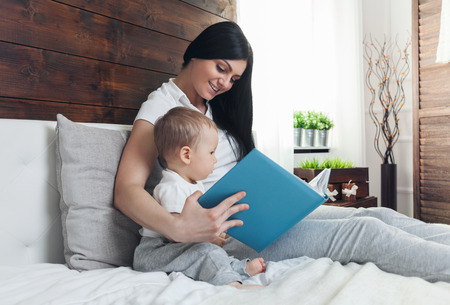 Child education. Happy mother with her toddler sitting on the bed and reading a book togetherの写真素材