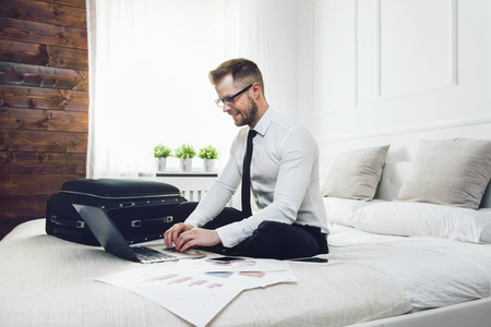 Young businessman on bed working with a laptop from his hotel roomの写真素材