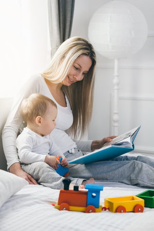 Child education. Happy mother with her toddler sitting on the bed and reading a book togetherの写真素材
