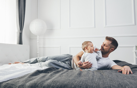 Happy father with his one year old son playing at home on the bedの写真素材