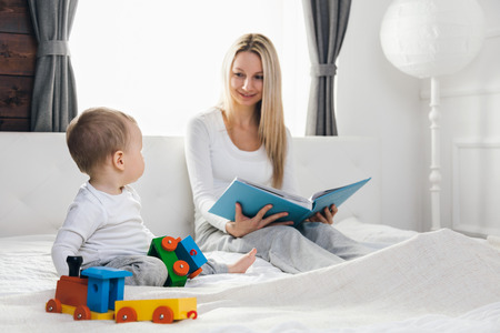Child education. Happy mother with her toddler sitting on the bed and reading a book togetherの写真素材
