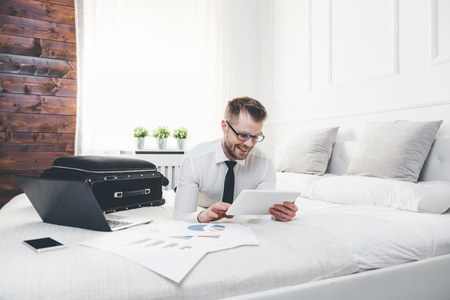Young businessman on bed working with a tablet laptop from his hotel roomの写真素材