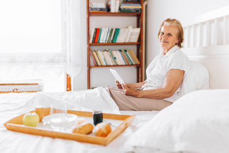 Happy elderly woman having breakfast in bed whilst using her tabletの写真素材