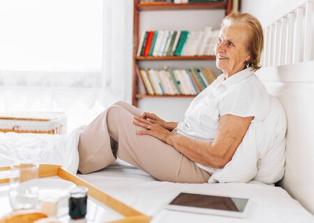Happy elderly woman having breakfast in bed whilst using her tabletの写真素材