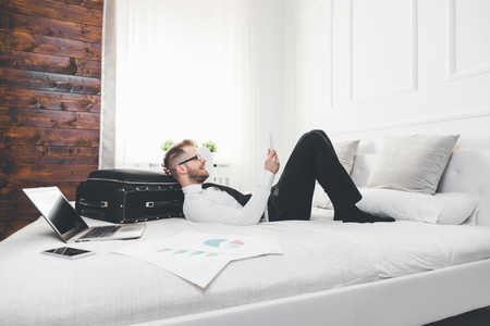 Young businessman on bed working with a tablet laptop from his hotel roomの写真素材