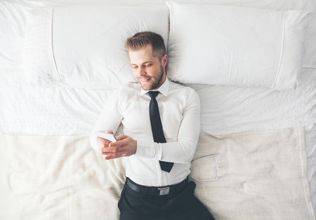 Top view. Young handsome businessman lying on the bed texting from his smartphoneの写真素材