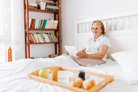 Happy elderly woman having breakfast in bed whilst using her tabletの写真素材
