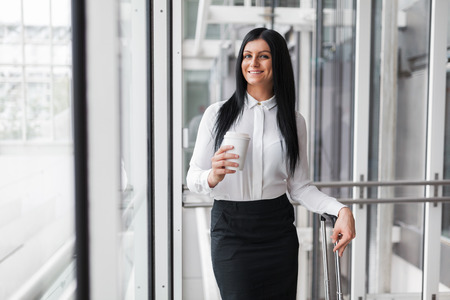 Successful confident young business woman with coffee and suitcase in an office settingの写真素材