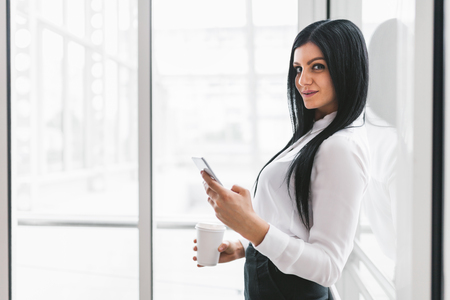 Successful confident young business woman with coffee and smartphone in an office settingの写真素材