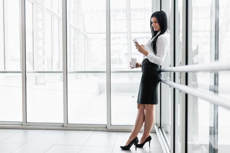Successful confident young business woman with coffee and smartphone in an office settingの写真素材