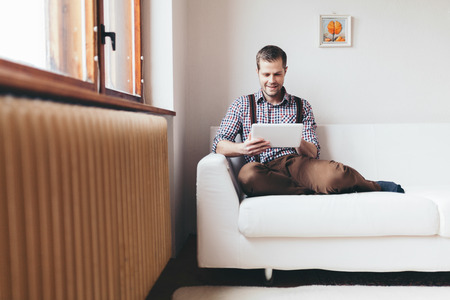 Handsome man sitting on sofa at home, using a digital tabletの写真素材