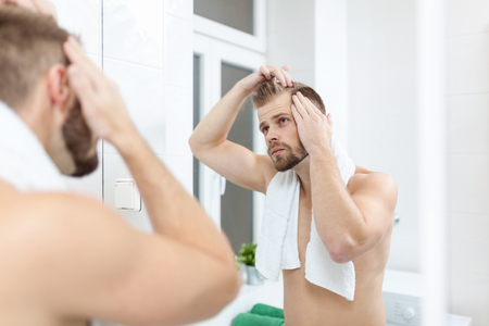 Handsome unshaven man looking into the mirror in bathroomの写真素材