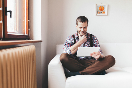 Handsome man sitting on sofa at home, using a digital tabletの写真素材