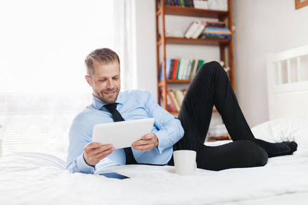 Young businessman on bed working with a tablet from his hotel roomの写真素材