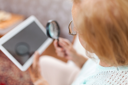 Elderly woman with glasses and loupe using a modern digital tabletの写真素材