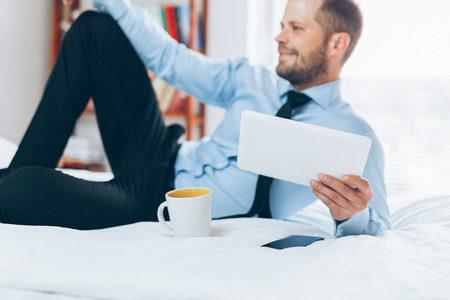 Young businessman on bed working with a tablet from his hotel roomの写真素材