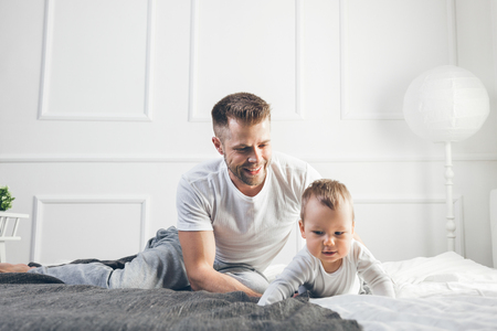 Happy father with his one year old son playing at home on the bedの写真素材