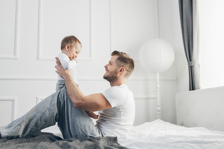 Happy father with his one year old son playing at home on the bedの写真素材