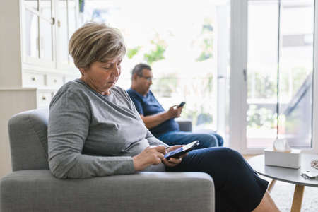 Senior couple sitting on couch and using their mobile phones at homeの写真素材