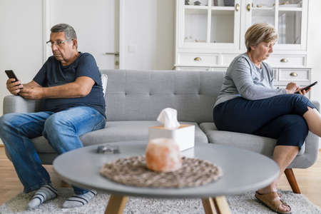Senior couple sitting on couch and using their mobile phones at homeの写真素材