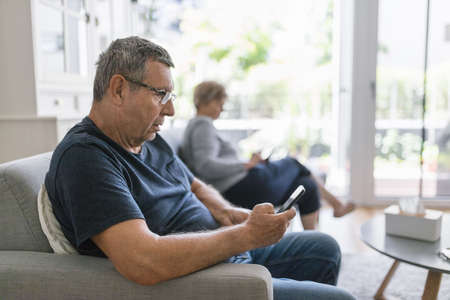 Senior couple sitting on couch and using their mobile phones at homeの写真素材