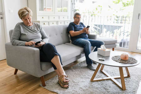 Senior couple sitting on couch and using their mobile phones at homeの写真素材