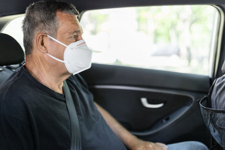 Senior in his 70s sitting at the back of car wearing a face mask for protection against corona virusの写真素材
