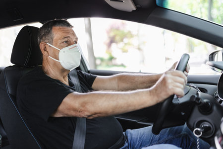 Senior in his 70s driving a car wearing face mask for protection against corona virusの写真素材