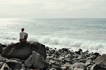 Isolated young woman thinking on a rock in the shoreline of the Atlantic Ocean coastの写真素材