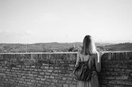 A young blonde woman is looking at the view beyond a brick wall (Corinaldo, Marche, Italy, Europe)の写真素材