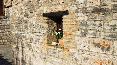 A small window under a wooden lintel decorated with a vase with white and violet flowers in a stone building of a medieval village (Marche, Italy, Europe)の写真素材