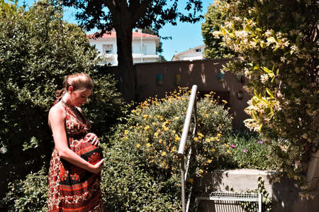 A pregnant blonde girl with a colored dress is stroking her belly in the garden on a spring day (Marche, Italy, Europe)の写真素材