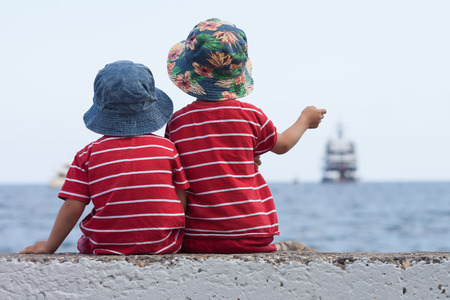 Two boys, contemplating a big ship in the sea, sitting on a wallの写真素材