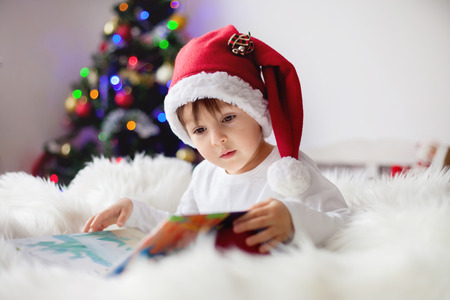 Cute adorable boy reading a book in front of the christmas tree, christmas timeの写真素材