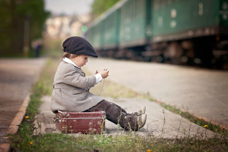 Boy, dressed in vintage coat and hat, with suitcase, on a railway station, steam trainの写真素材