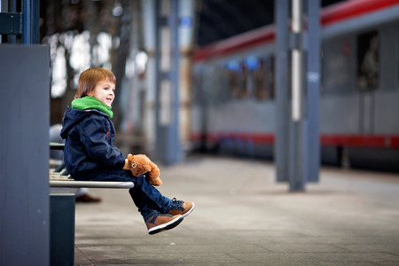 Cute boy, sitting on a bench with teddy bear, looking at a train, leaving the railway stationの写真素材