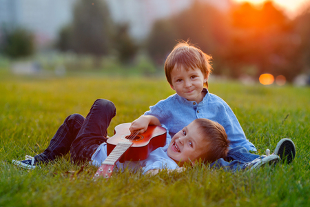 Two adorable boys, sitting on the grass, playing guitar on sunsetの写真素材