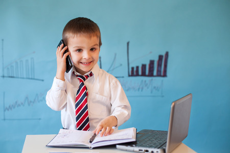 Smart little boy, working on computer, taking notes and speaking on the phoneの写真素材