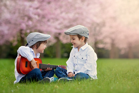 Two adorable caucasian boys in a blooming cherry tree garden, playing guitar in the afternoonの写真素材