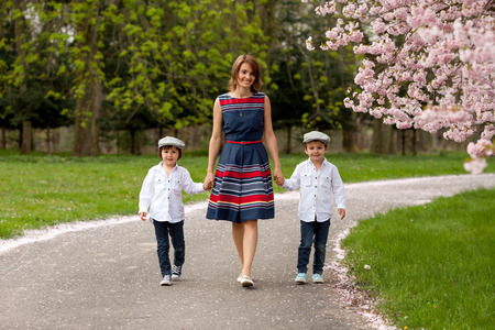 Beautiful portrait of mother and her two children in a cherry blossom tree garden, waking down the alley, holding hands and looking at the cameraの写真素材