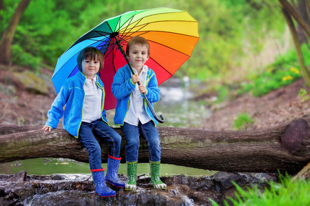 Two adorable little boy, brothers, sitting on a wooden trunk on a pond in a rainy day with colorful umbrella, boots and casual clothing, smiling at the cameraの写真素材