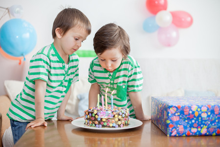 Beautiful adorable four year old boy and his brother in green shirts, celebrating his birthday, blowing candles on homemade baked cake, indoor. Birthday party for kidsの写真素材