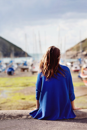 Young woman, sitting at the harbor on the quay with her back to the camera looking out at the moored boats, shallow dofの写真素材