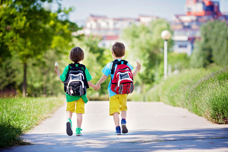 Two adorable boys in colorful clothes and backpacks, walking away, holding and eating ice cream on a sunny summer afternoon, warm day, casual clothingの写真素材