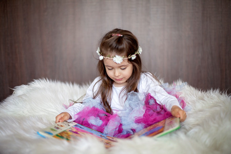 Sweet little girl, sitting in big chair with white fur, reading a book, wearing tutu skirt and headbandの写真素材