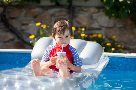Little boy in a big swimming pool, drinking juice in a hot summer day, sitting on a floating bedの写真素材