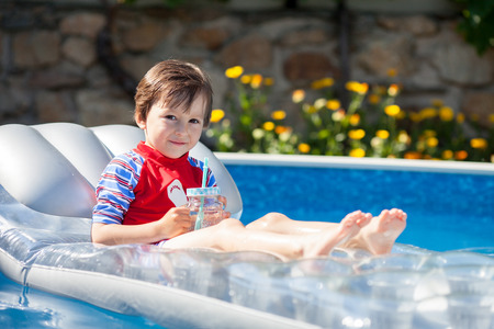 Little boy in a big swimming pool, drinking juice in a hot summer day, sitting on a floating bedの写真素材
