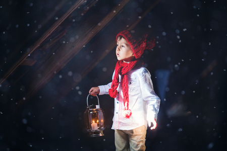 Sweet boy, holding a lantern, looking at a light coming through a window, standing in the snow, outdoorsの写真素材